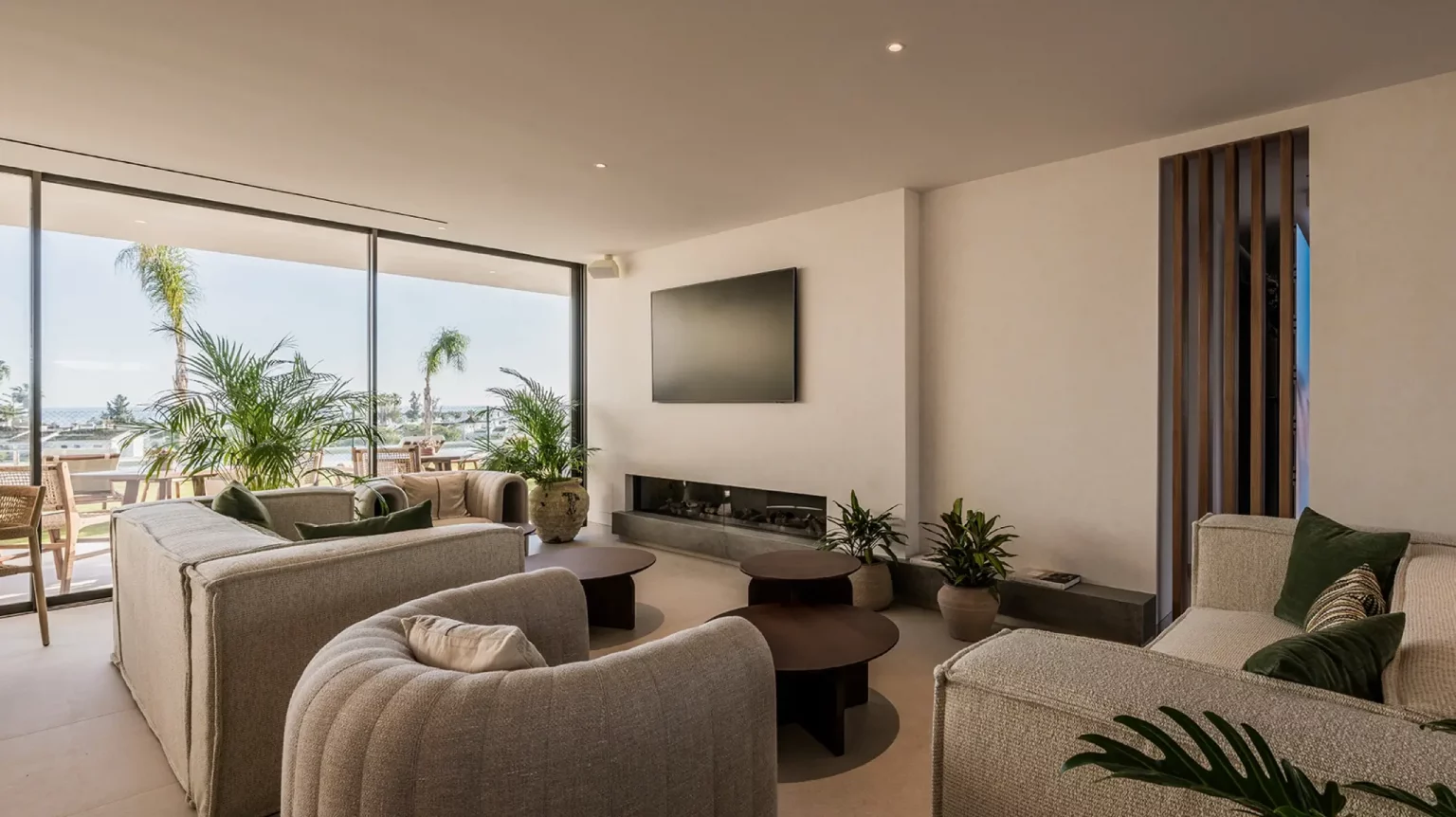 Bright modern living room with beige sectional sofas, potted plants, a wall-mounted TV, and a glass wall overlooking a patio ground.