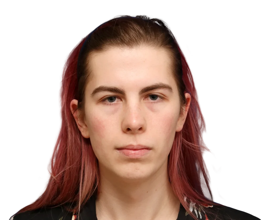 Close-up portrait of Elléonore with pale skin and pink-red hair, looking straight ahead with a neutral expression against a black background.