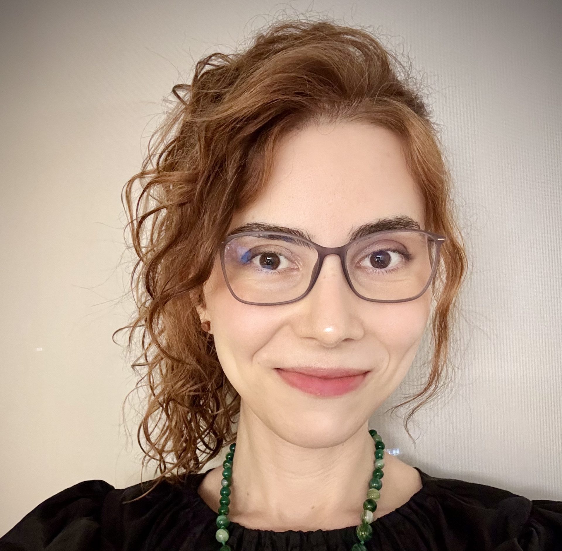 Portrait of a woman with red curly hair, wearing gray rectangular glasses, a green beaded necklace, and a black top, smiling at the camera.