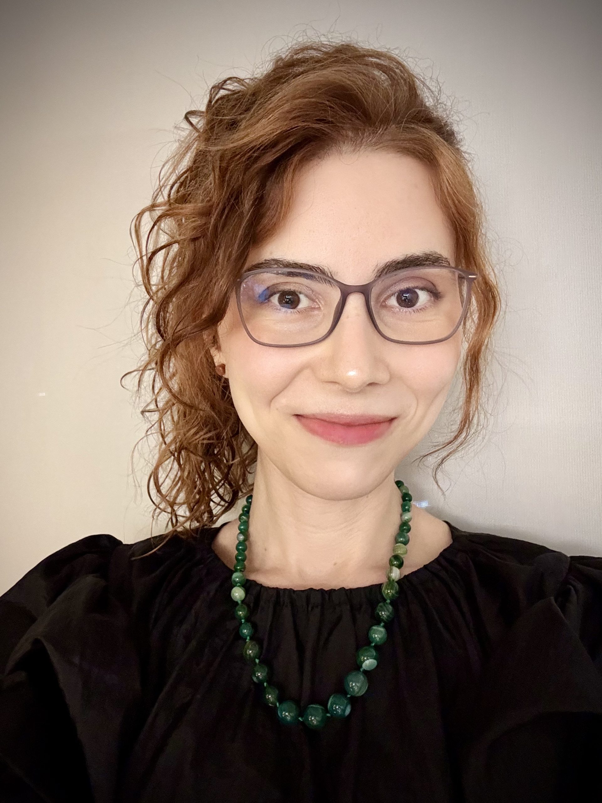 Portrait of a woman with red curly hair, wearing gray rectangular glasses, a green beaded necklace, and a black top, smiling at the camera.