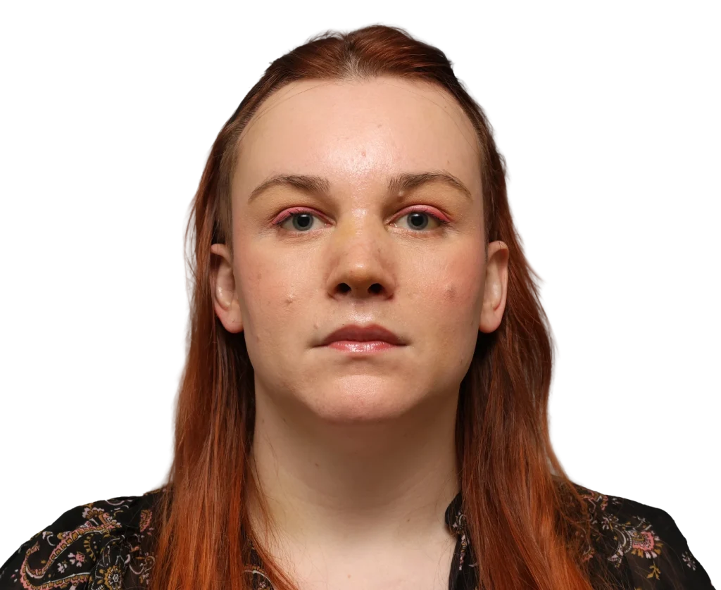 Front-facing portrait of a Mia with long red hair and pink eyeshadow, wearing a black floral blouse against a dark background.